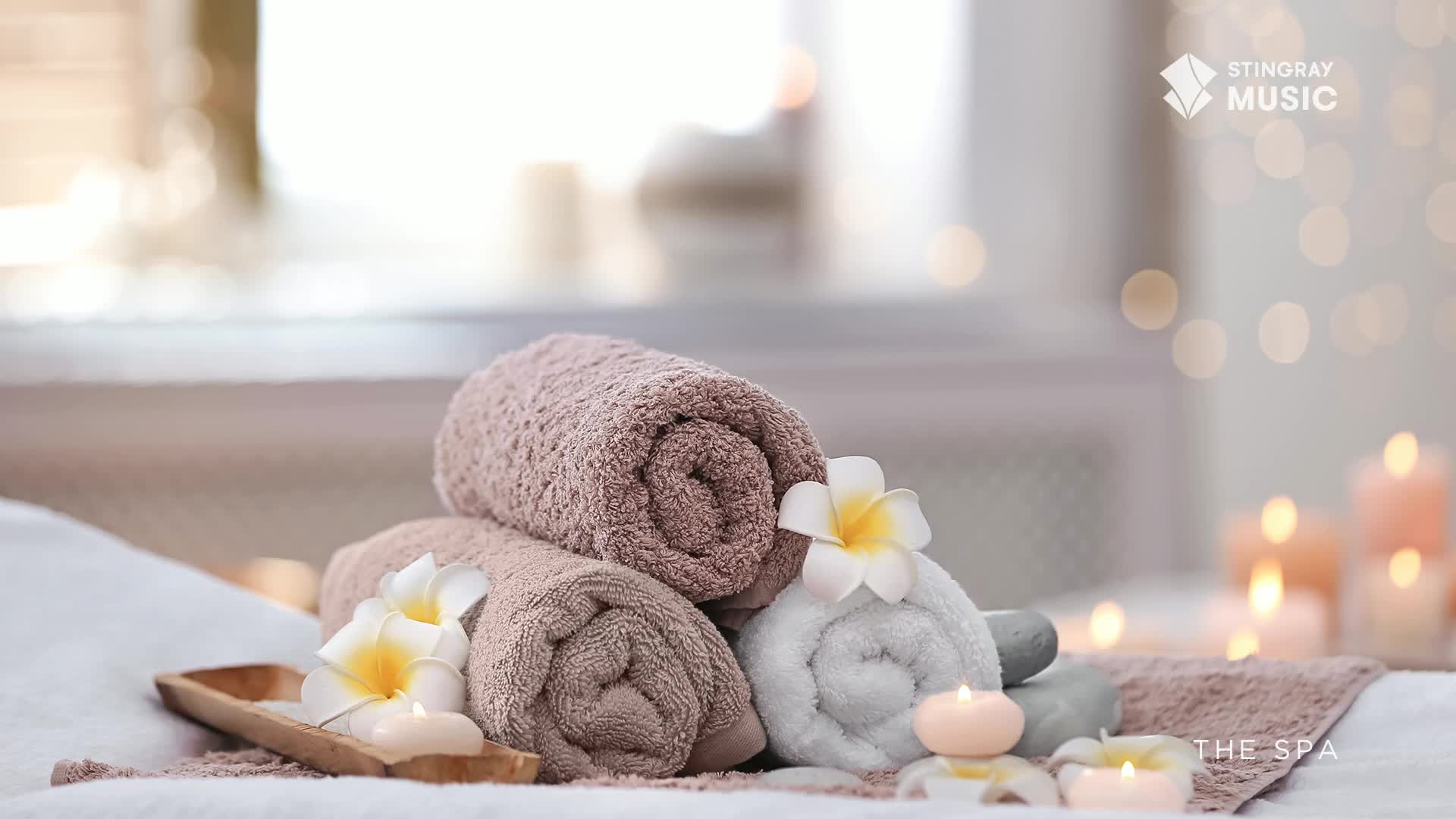 Soft, rolled towels rest on a spa table, adorned with delicate flowers and lit candles. The scene, bathed in a warm glow, evokes a sense of calm and relaxation.
