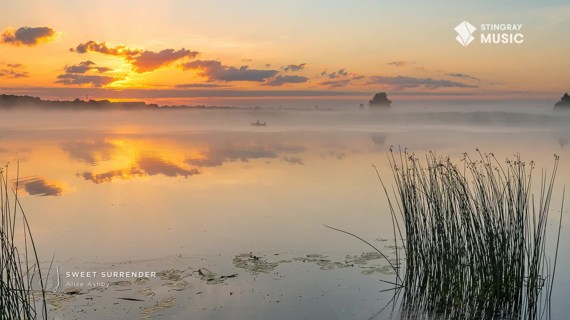 The sun is rising, painting the sky and clouds with warm hues that reflect in the still water. A thin layer of mist hangs over the lake, softening the distant shoreline and obscuring a small boat.
