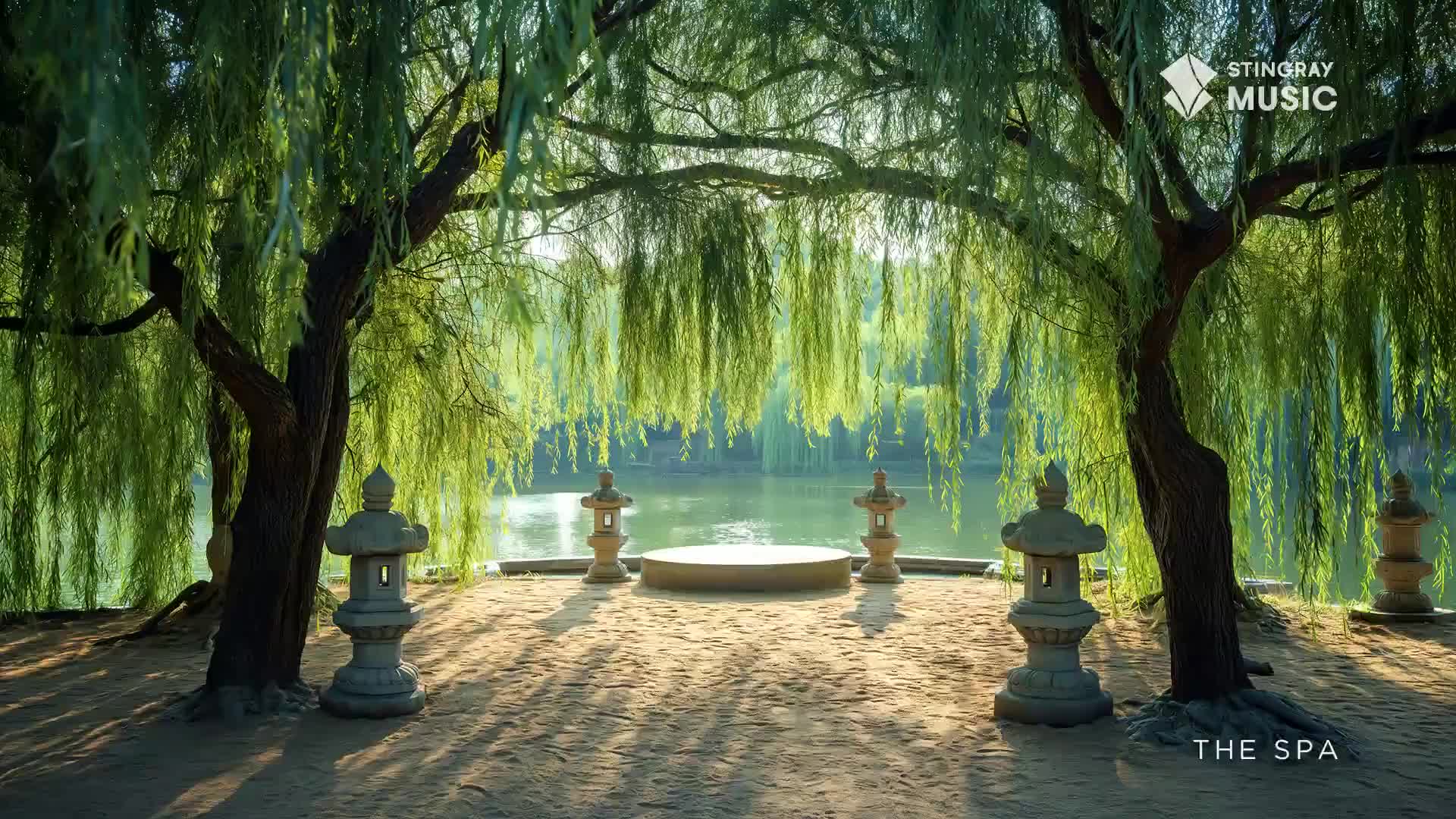 Sunlight dapples the ground beneath the weeping willow trees, casting long shadows across the stone lanterns. Beyond the trees, a small, circular platform overlooks a tranquil lake, perfect for a Stingray The Spa experience.
