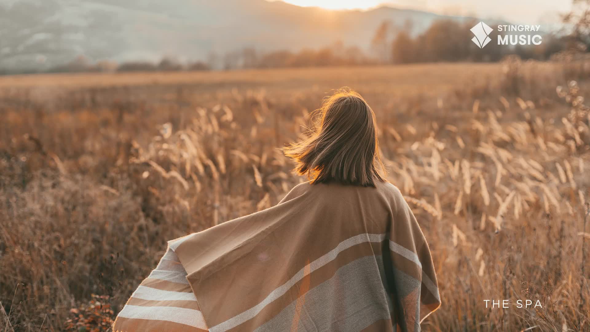 A woman walks through a field of tall, dry grass, a blanket draped over her shoulders. The sun sets in the distance, casting a warm glow over the scene, which feels like a moment of peace from Stingray The Spa in Canada.
