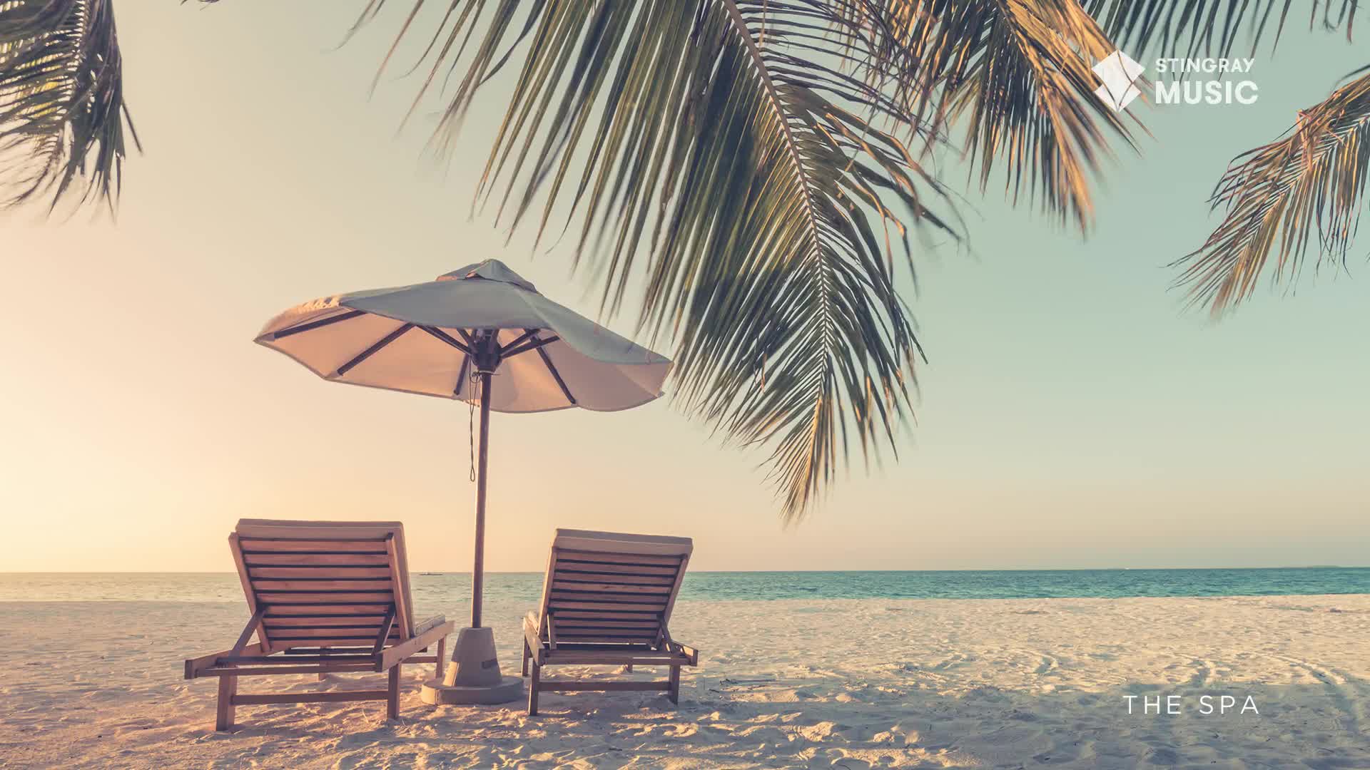 Underneath a gently swaying palm frond, two wooden beach chairs sit empty, facing the ocean. The sunshade of an umbrella offers a bit of respite from the warm glow of the Canadian sky.
