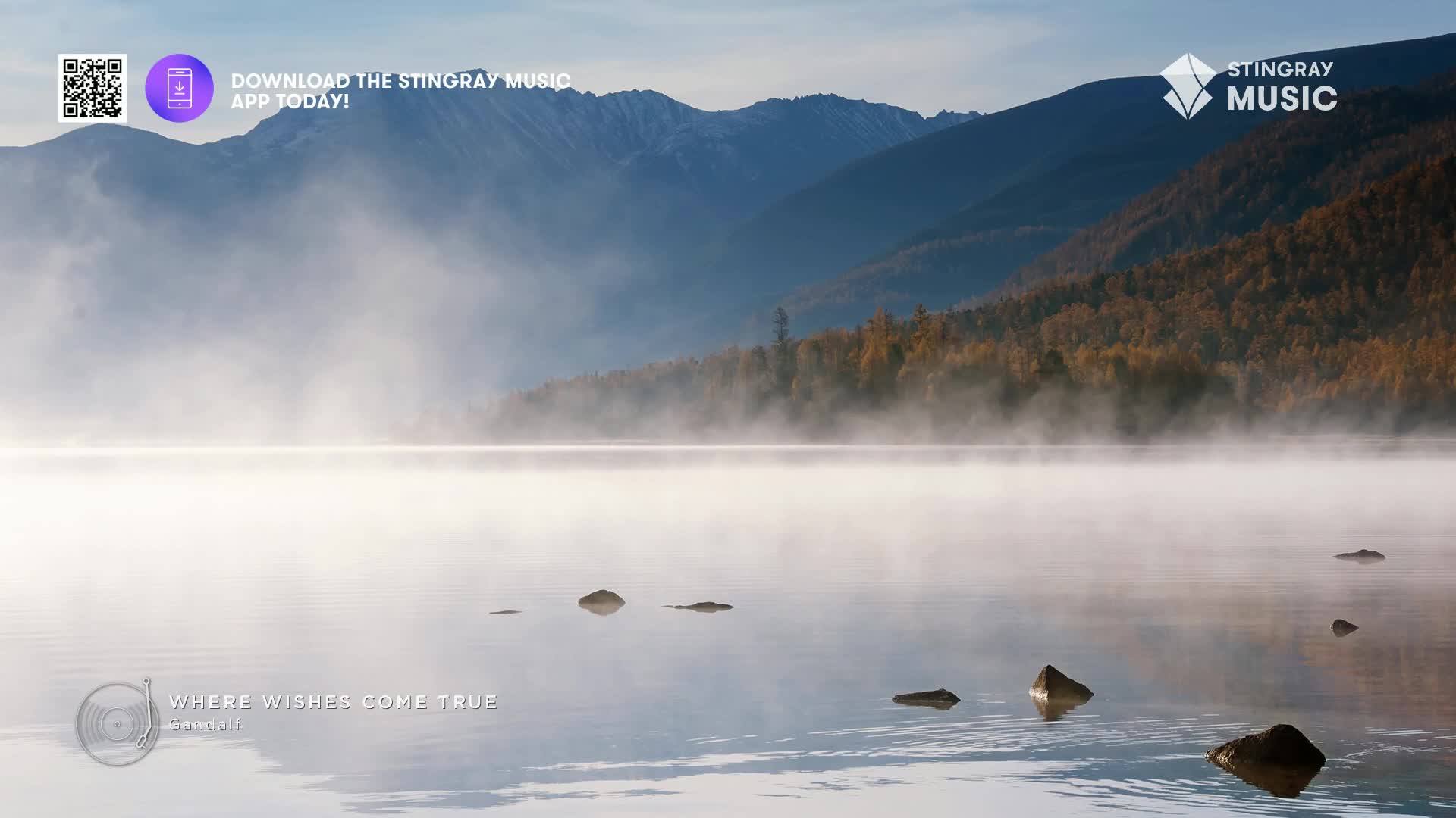 Mist rises from the still water of a Canadian lake, its surface dotted with dark rocks. The distant mountains, shrouded in a soft haze, frame the tranquil scene.