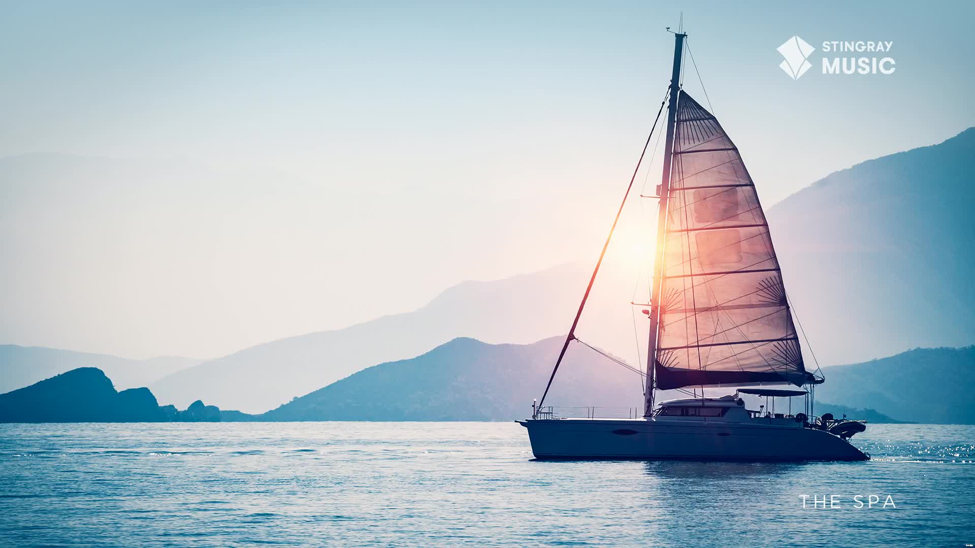 A catamaran glides across calm water, its sails catching the low sun. Distant, layered mountains rise behind it, hinting at a Canadian coastline.