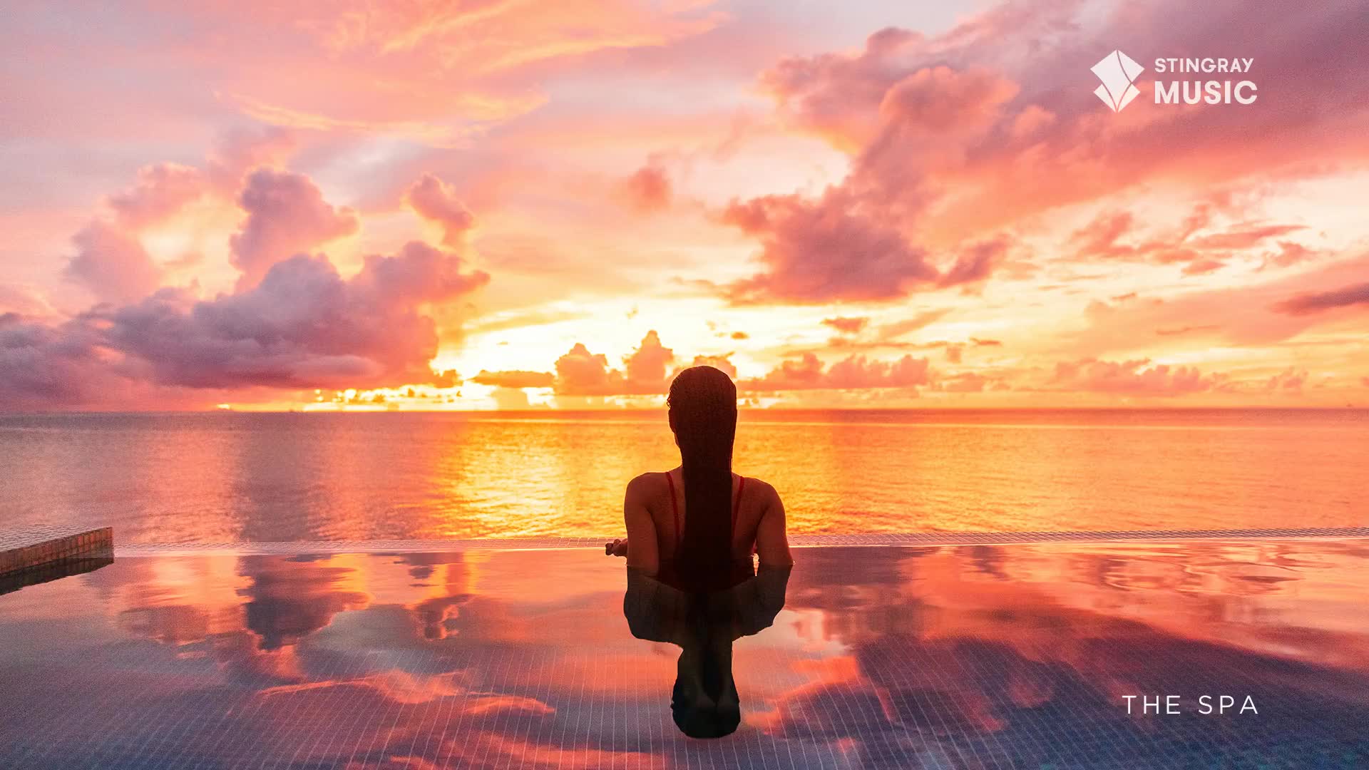 The sun dips below the horizon, painting the sky in fiery oranges and soft pinks. A woman sits in an infinity pool, her reflection shimmering on the calm water.