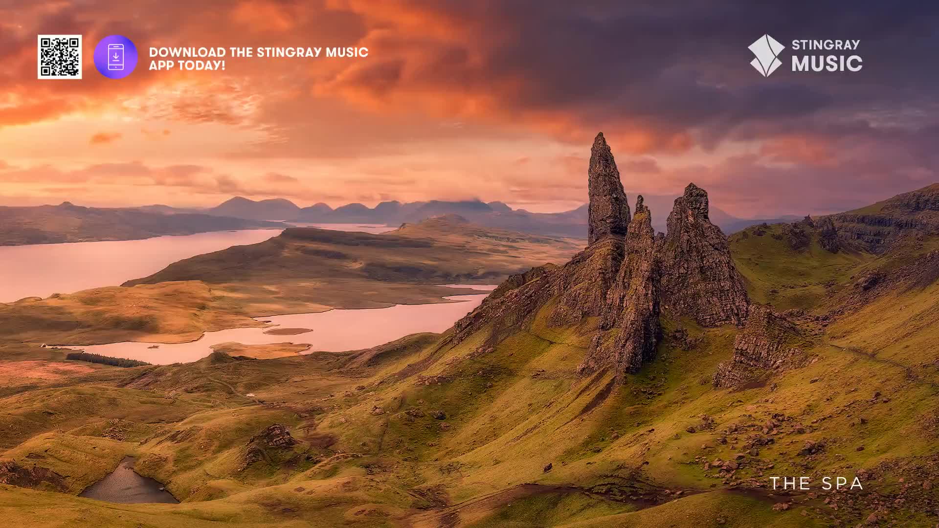The sky above the rugged, green hills is painted with vibrant orange and purple hues. A steep, rocky spire stands sentinel against the dramatic sunset.