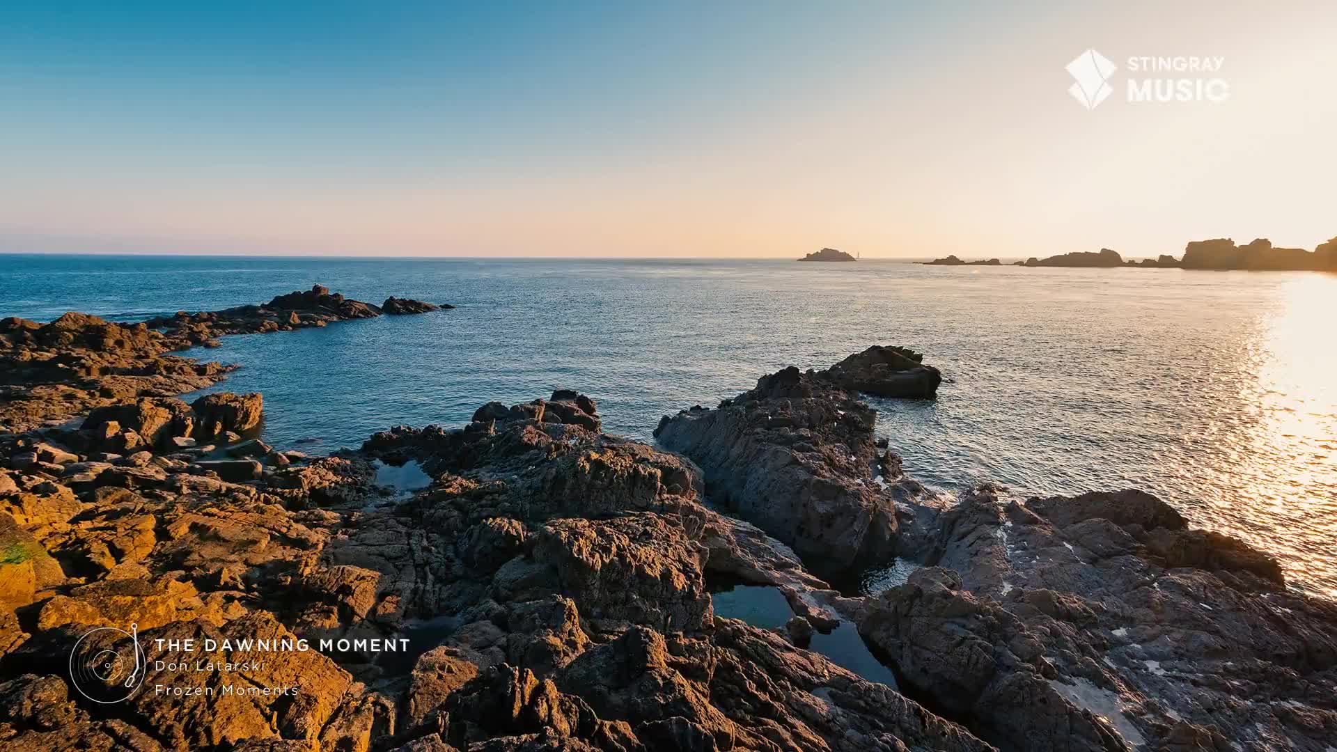 The sun is just beginning to touch the horizon, casting a warm glow across the rugged Canadian coastline. Gentle waves lap against the dark, weathered rocks, reflecting the soft light of dawn.