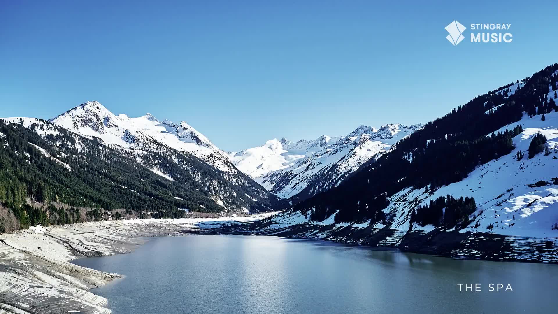 The vast, still water of the reservoir reflects the snow-capped peaks of the Canadian Rockies. Dark evergreen trees cling to the steep slopes, their shadows stark against the bright snow.