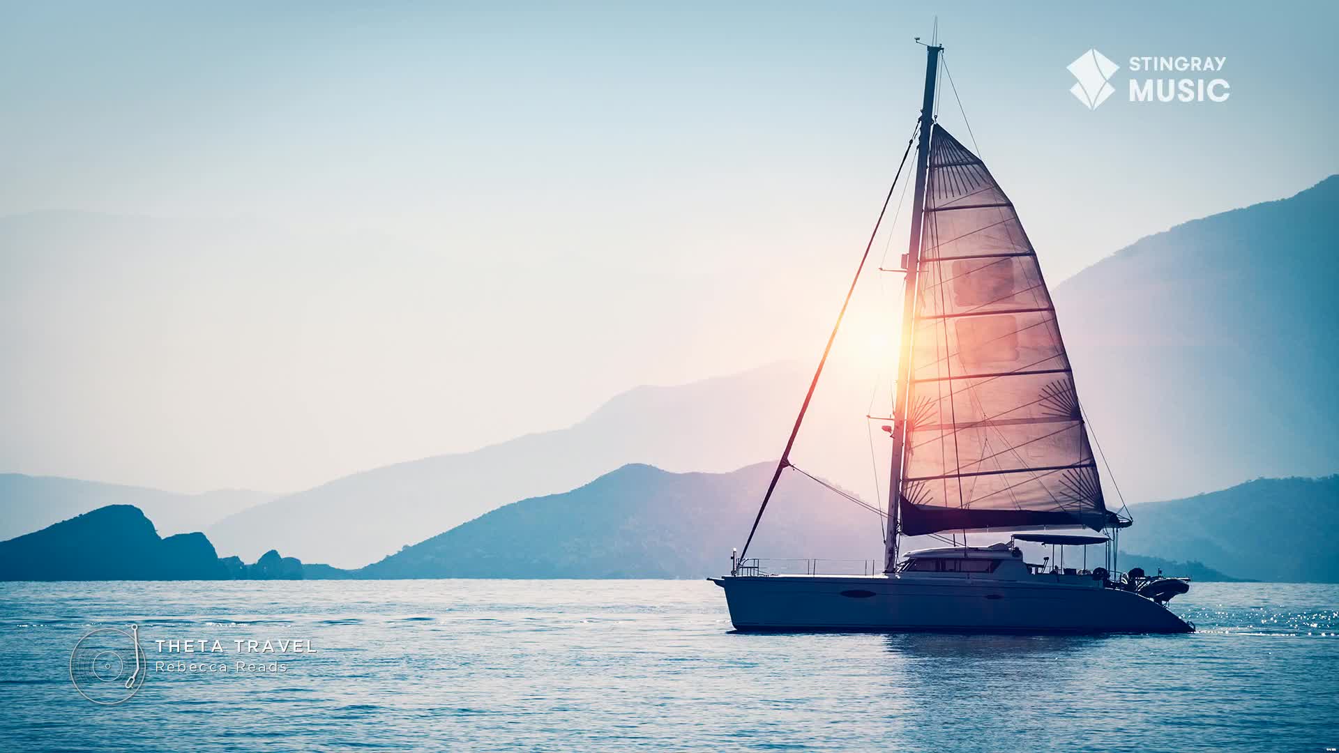 A catamaran glides across the water, its sails catching the late afternoon sun. Distant, hazy mountains rise in layers behind the boat, their peaks softened by the light.