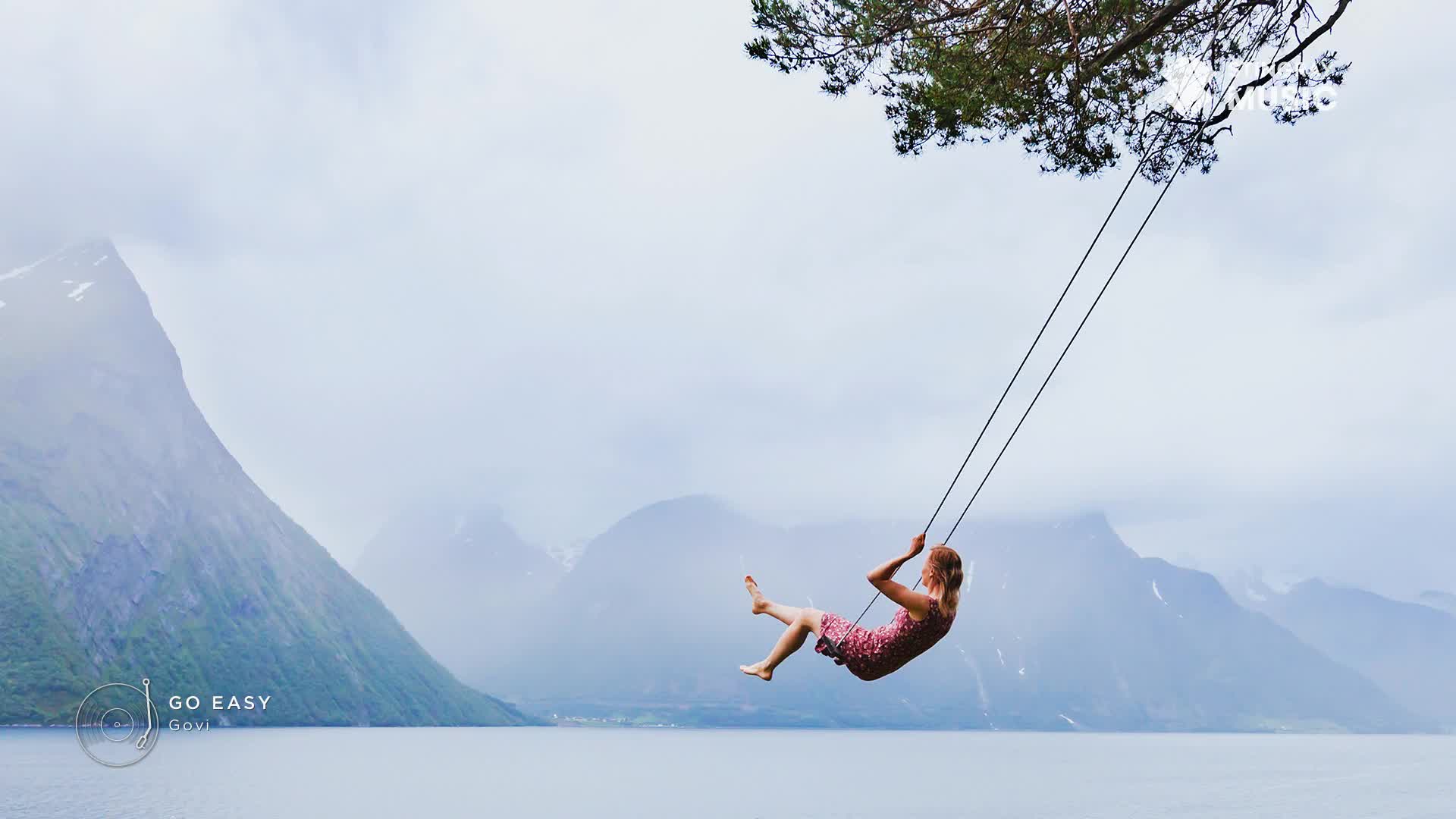 A young woman in a pink dress swings out over a calm lake, her bare feet reaching for the sky. Towering, mist-shrouded mountains rise behind her, a classic Canadian landscape.