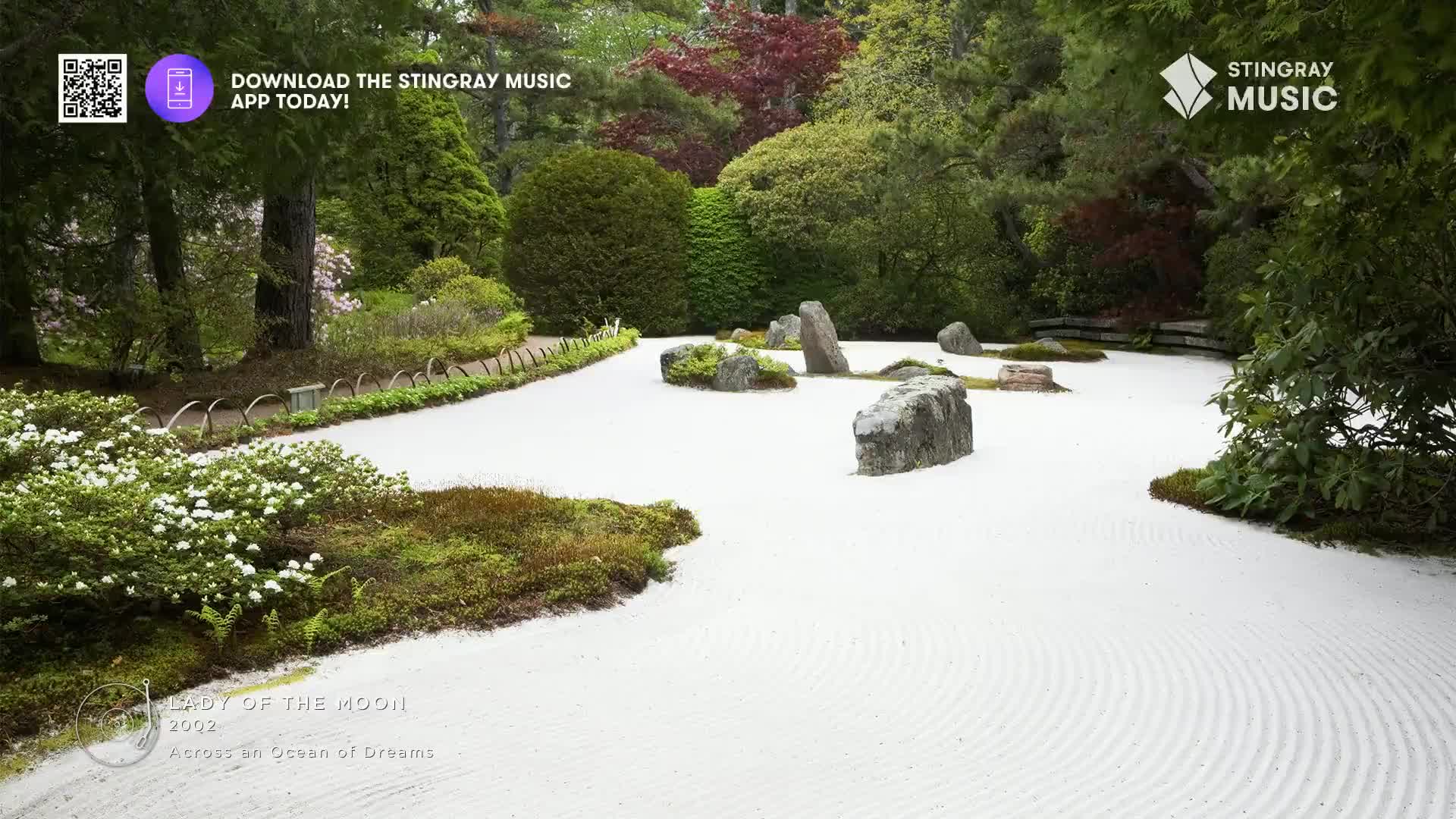The raked white sand of this Japanese garden in Canada creates a serene backdrop for the carefully placed rocks. Lush green foliage and delicate white blossoms frame the tranquil scene.