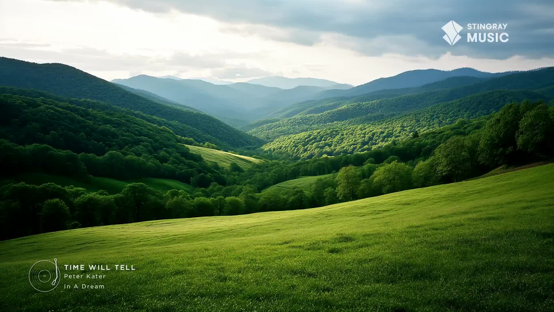 Rolling green hills stretch out before me, leading into a valley carved between forested mountains. The sky above is a mix of soft clouds and bright sunlight, illuminating the lush Canadian landscape.