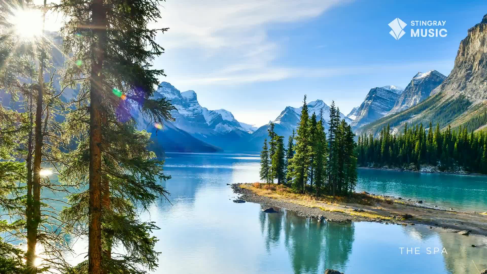 Sunlight streams through the pine branches, illuminating the vast, tranquil lake. Jagged, snow-capped mountains rise majestically in the distance, framing the serene Canadian landscape.
