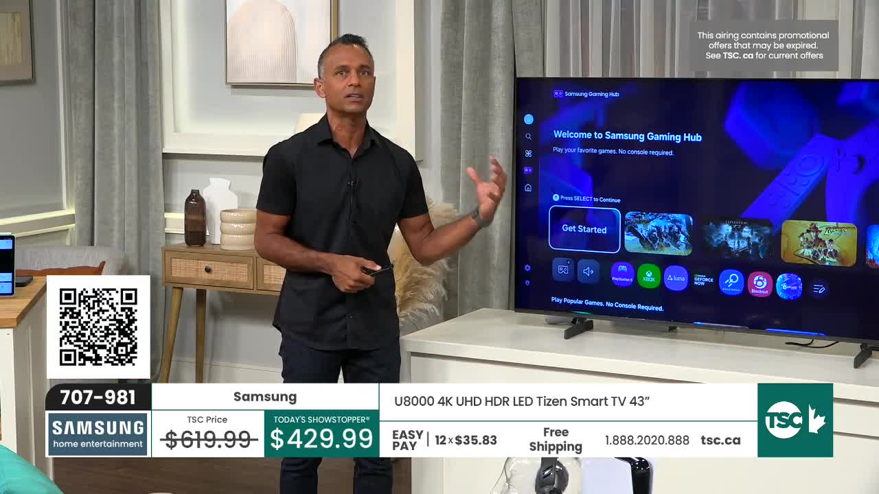 A man in a black shirt gestures toward a large television screen displaying a gaming hub interface. The screen is flanked by a TSC Canada logo and a price tag for a Samsung TV.

