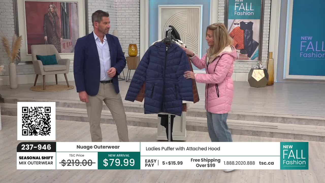 A woman in a pink jacket holds up a navy blue puffer coat with brown accents, while a man in a blazer looks on. They're on a TSC set, likely in Canada, showcasing the outerwear for the fall season.
