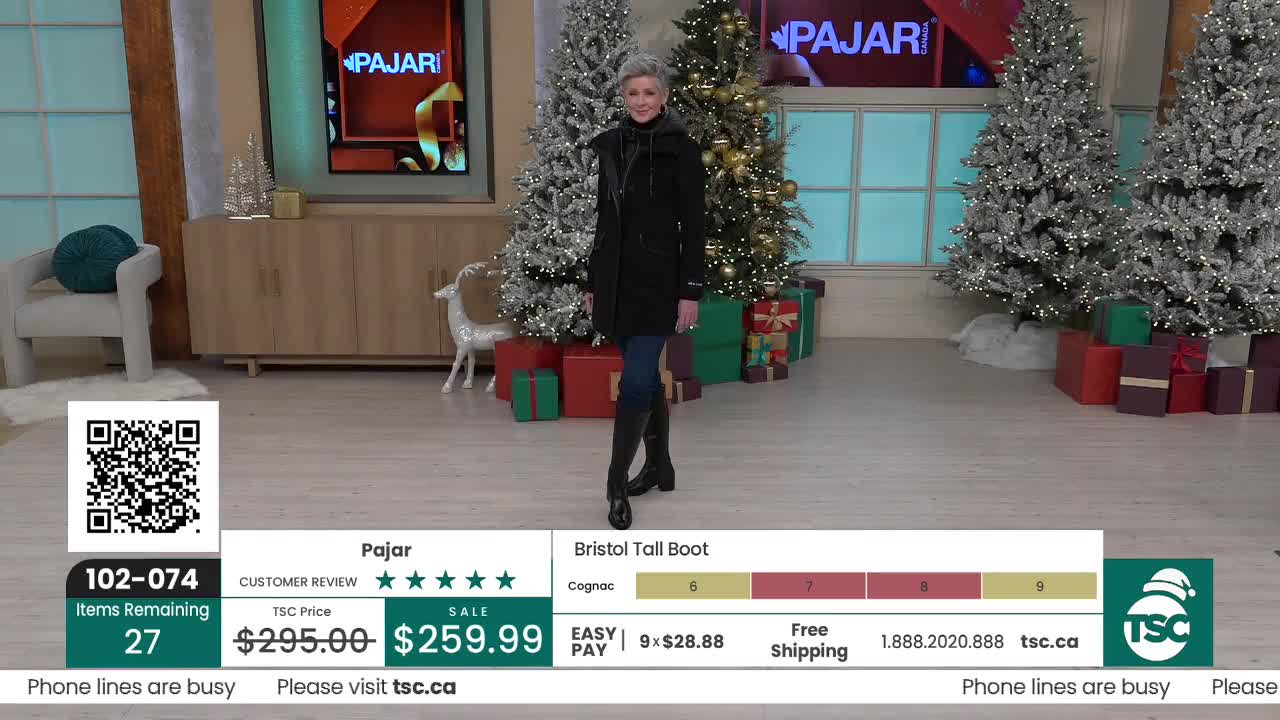 A woman in a black coat and boots is center stage, smiling as she models the footwear. Behind her, Christmas trees and wrapped gifts decorate the set of a TSC Canada broadcast.
