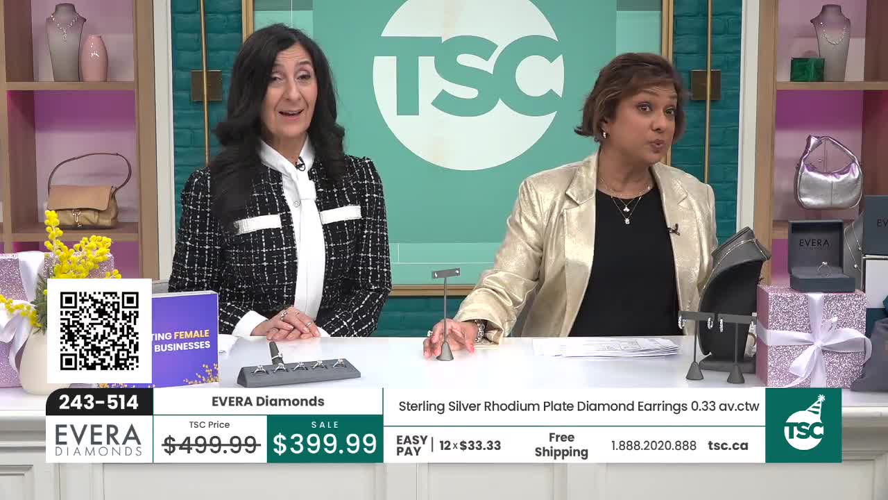 Two women stand behind a table displaying jewelry. The woman on the right holds up a necklace and earrings, explaining their details.