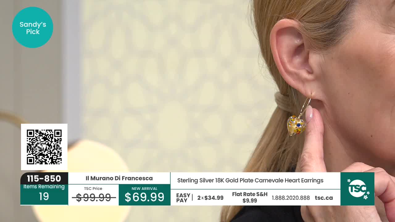 A woman's finger gently touches a dangling heart-shaped earring, its glass catching the light. The earring, a "Sandy's Pick" from TSC, features swirling colours within its golden form.