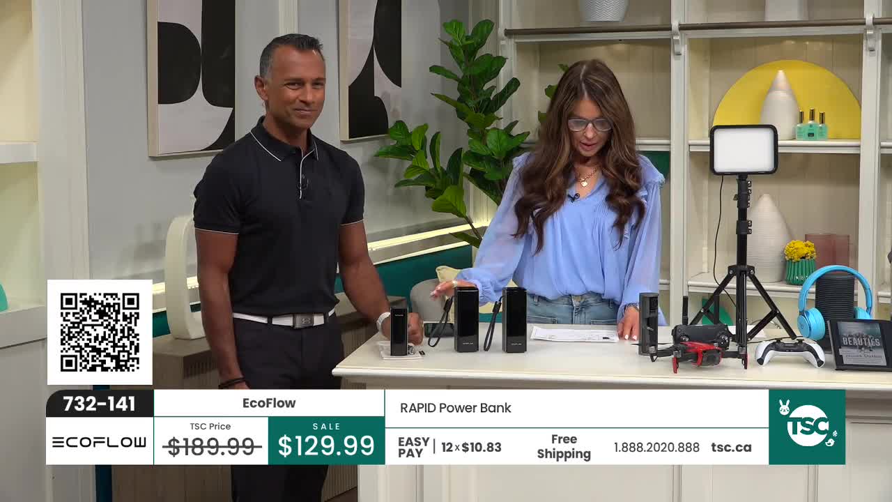 A man and woman stand behind a table displaying various electronic items, including power banks and what looks like a small drone. The woman gestures towards the items as she speaks, while the man smiles and looks on. A man and woman stand behind a table displaying various electronic items, including power banks and what looks like a small drone. The woman gestures towards the items as she speaks, while the man smiles and looks on.
