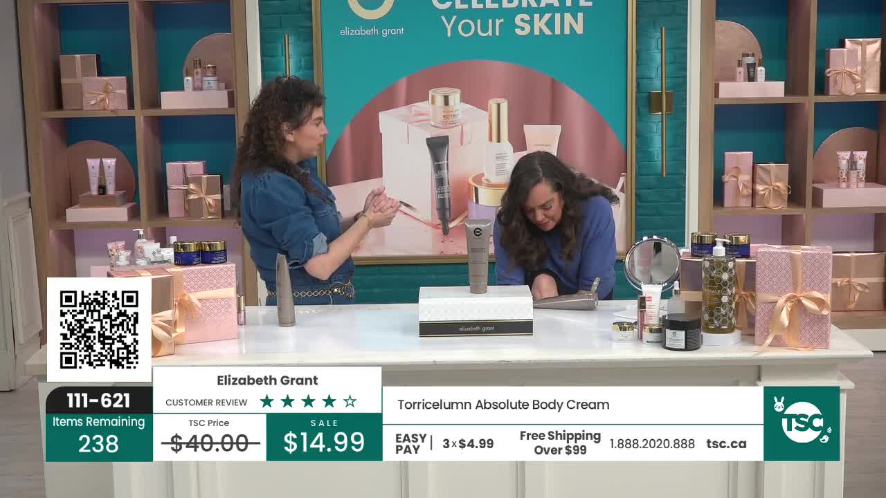 A woman in a denim shirt gestures with her hands as another woman in a blue sweater leans over a table. On the table, a tube of Torricelumn Absolute Body Cream sits in front of a white box.