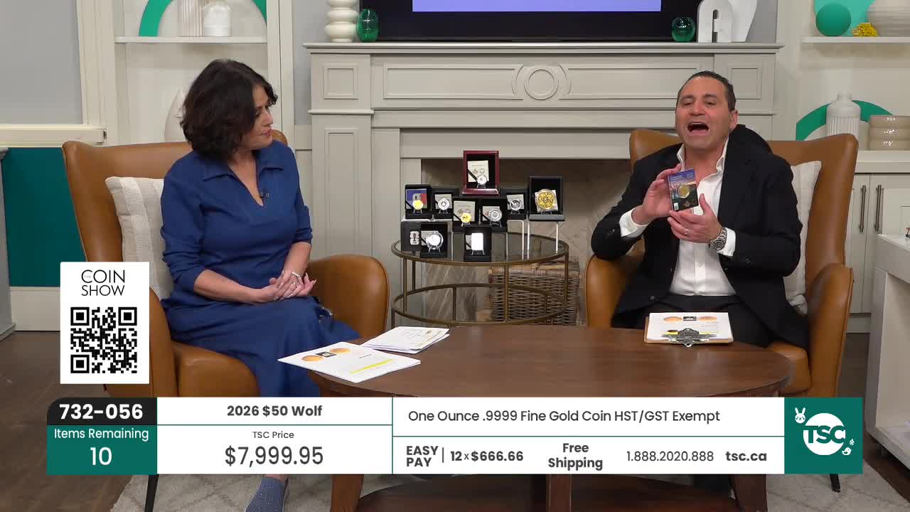 A man in a suit enthusiastically holds up a small, colourful package, gesturing with his free hand. Beside him, a woman in a blue dress sits calmly, her hands clasped in her lap.