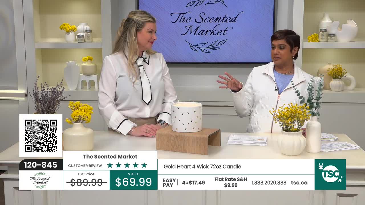 A woman in a white blouse with a black tie gestures animatedly as she speaks. Beside her, a large white candle with black hearts sits on a wooden stand.