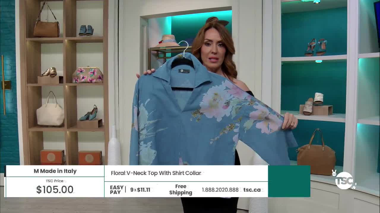A woman holds up a flowing blue top with a large floral print, showcasing it for the camera. Shelves behind her display handbags and shoes, typical of a shopping channel presentation.