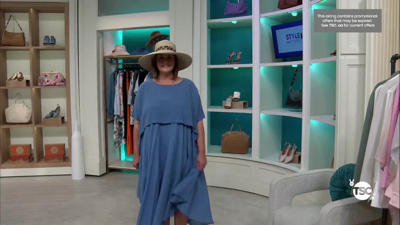 A woman in a wide-brimmed hat and a flowing blue dress is presenting on TSC. She gestures with her right hand, her dress pooling around her feet.