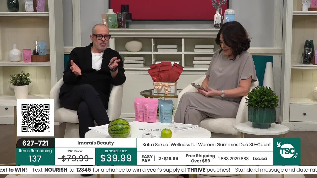 A man in a black jacket gestures with his hands while speaking, and a woman in a grey dress looks at her phone. A small watermelon and two pouches of gummies sit on a coffee table between them.