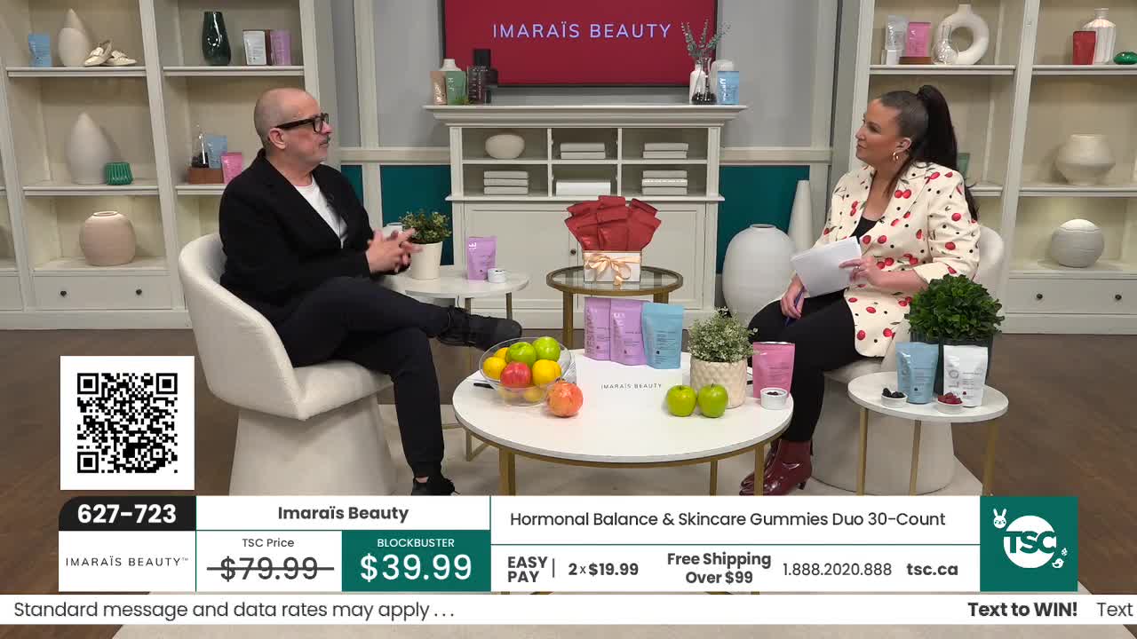 A man in a black blazer and a woman in a cherry-print jacket are seated, discussing products. The woman holds a piece of paper as they chat on what looks like a set for a Canadian shopping channel.