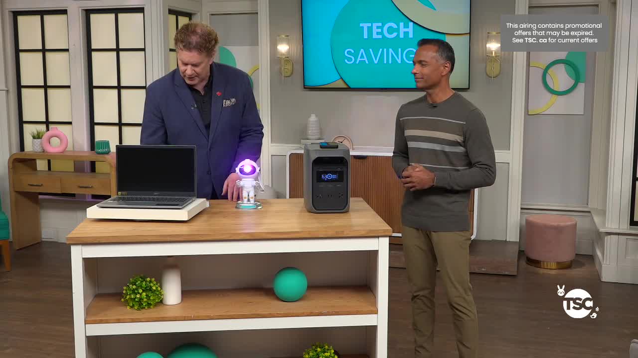A man in a blue blazer gestures towards a small, illuminated robot on a table. Beside it, a portable power station sits ready for demonstration.