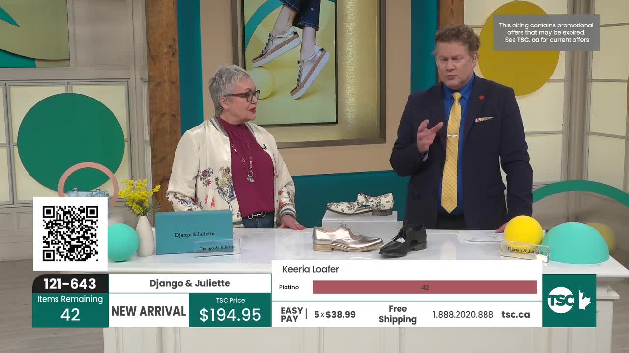 A woman in a floral jacket and a man in a suit stand behind a display of shoes. The man gestures as he speaks, pointing to a pair of black loafers.