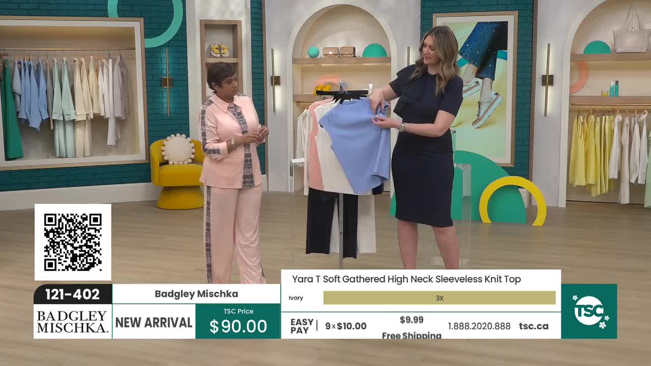 A woman in a navy dress holds up a light blue knit top for display. Beside her, another woman stands in a matching pink pantsuit, looking towards the camera. A woman in a navy dress holds up a light blue knit top for display. Beside her, another woman stands in a matching pink pantsuit, looking towards the camera.