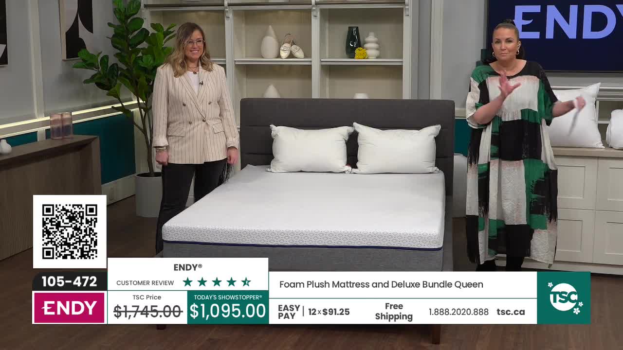 Two women stand on either side of a queen-sized mattress on a bed frame. The woman on the left wears a pinstripe blazer, and the one on the right has on a colorful, abstract-patterned dress.