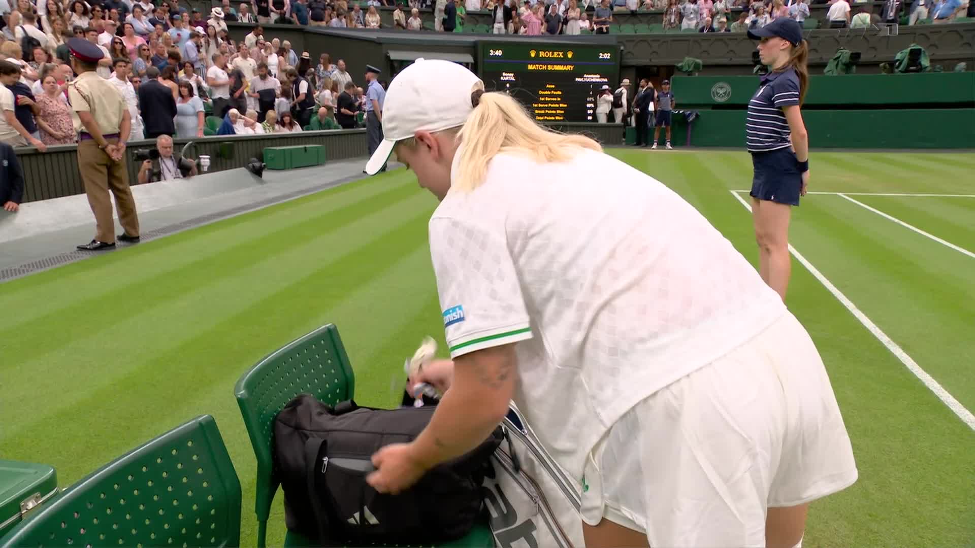 The player is bent over, rummaging through her black bag, a white towel in her hand. A lineswoman stands at attention on the court, watching the action.
