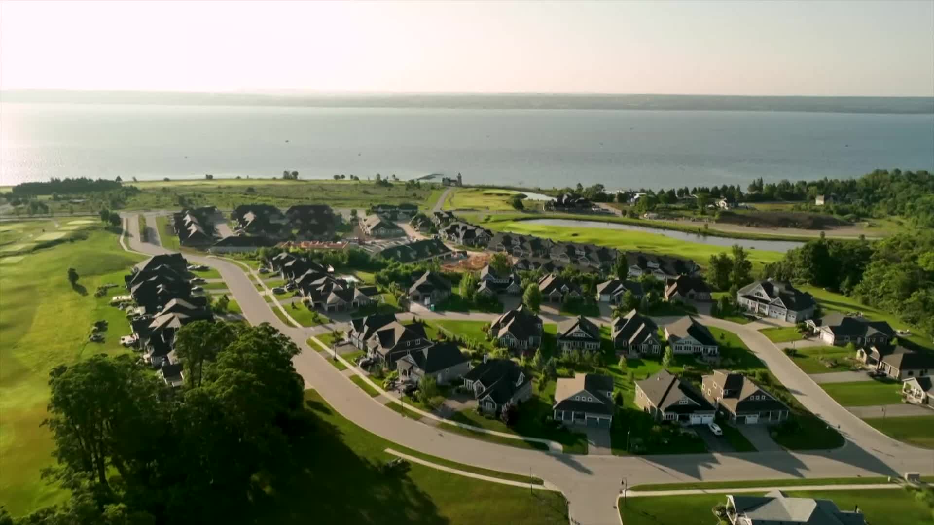 The camera sweeps across a residential area, with rows of houses nestled against a golf course. Beyond the green, a large body of water shimmers in the sunlight.
