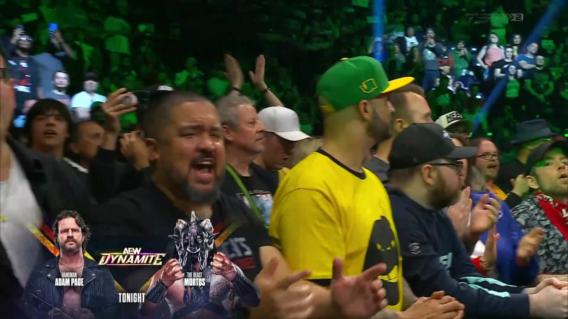 The crowd is amped up, with many people recording the action on their phones. A man in a yellow shirt claps, while the guy next to him wears a green hat and watches intently.
