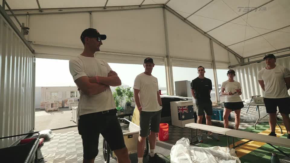 Under a white tent, a group of people in matching shirts and caps stand around, listening. The TSN2 logo is visible in the corner, and the scene suggests a briefing or a post-race debrief.
