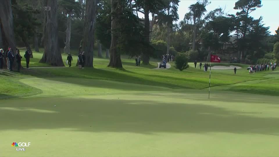A small golf cart putters along a path, while a group of people follow. The flag waves in the distance, marking the hole on the green.
