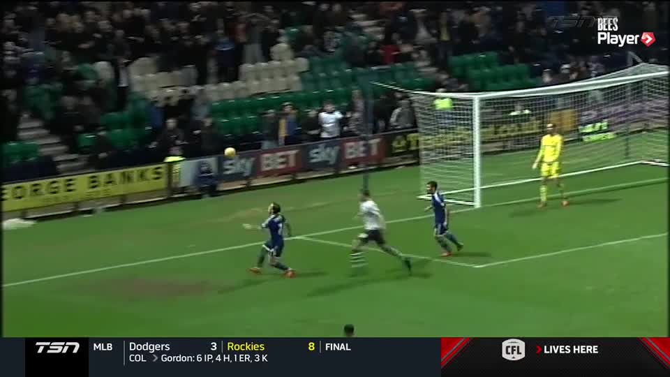 The ball hangs in the air, arcing toward the goal as a player in blue sprints to catch up. The goalie, in yellow, stands ready, watching the trajectory.
