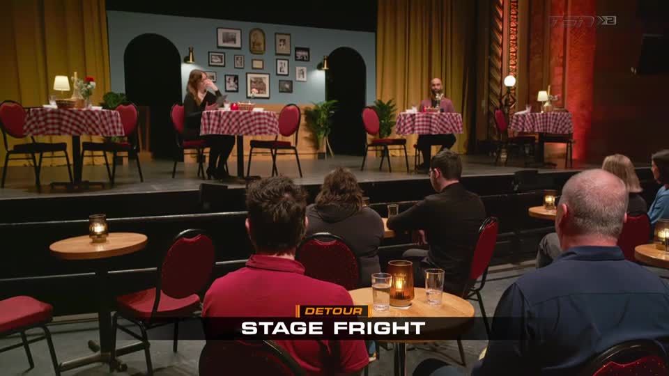 On a stage, two people sit at separate tables, each covered with a red-and-white checkered cloth. The audience, seated at small tables, watches the performance, drinks in hand.
