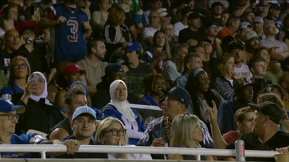 The crowd is packed, and many are looking toward the field. A woman in a hijab is holding up her phone, while a man in a baseball cap points up with his index finger.
