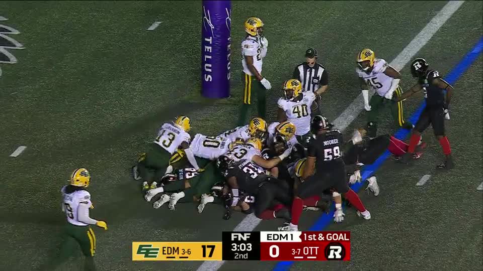 A pile of players in green and black shirts is crammed together near the goal line. The referee watches closely as a player in black tries to push through the scrum.
