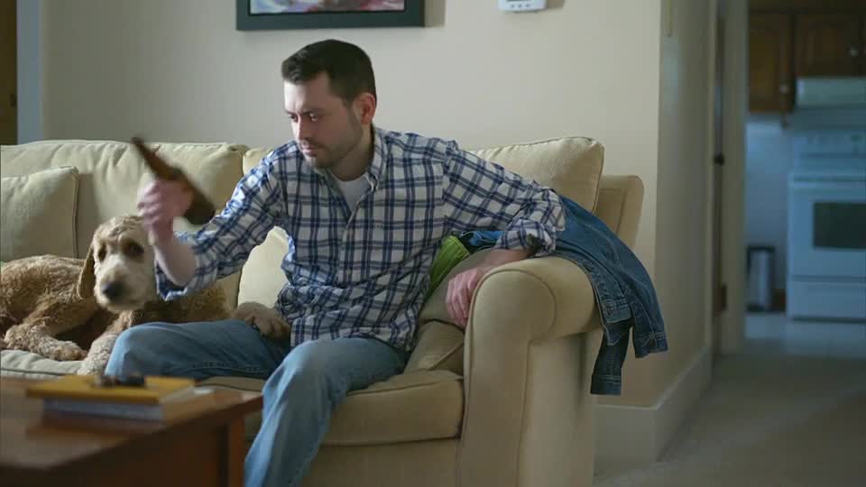 A man, beer bottle in hand, gestures toward a dog lounging on the couch beside him. He's likely watching something on TSN2, judging by his focus.
