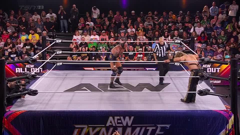 The wrestler in the center is hunched over, catching his breath, while the other leans against the ropes. A referee in black and white stripes hovers nearby, watching the action unfold on TSN2 in Canada.
