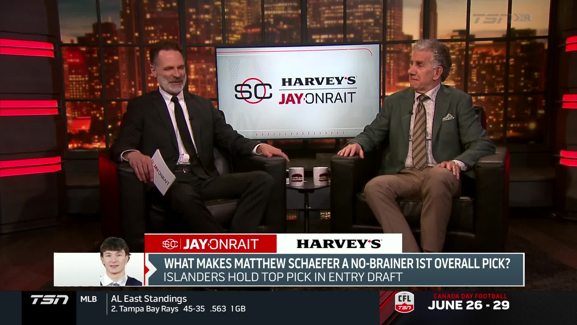 Two men in suits sit in armchairs, facing each other in a studio. The "SC Jay Onrait" logo is visible on the screen behind them, and the bottom banner mentions the CFL.
