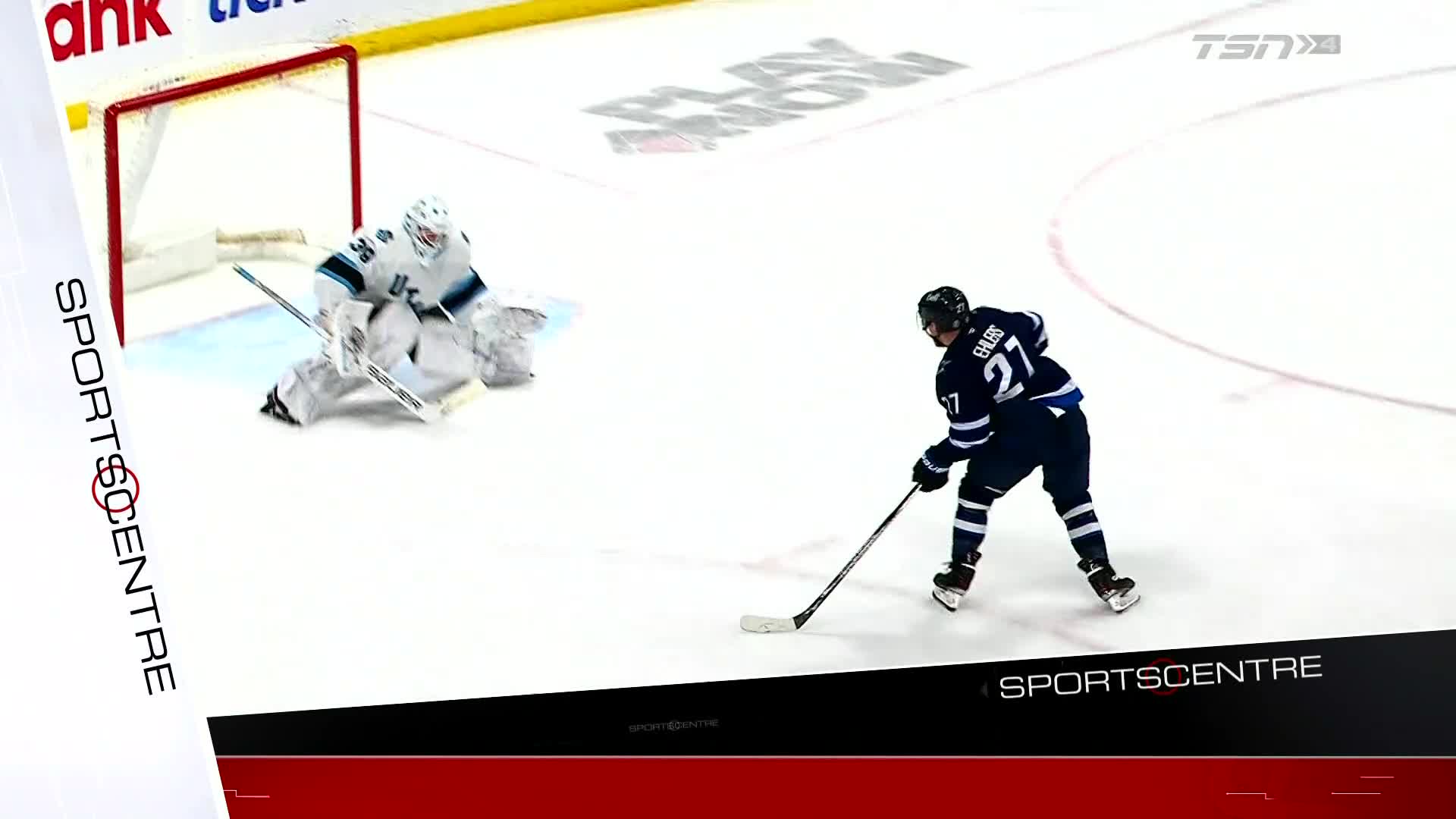 The goalie is down, poised to block the shot. A player in a blue jersey, number 27, skates toward the net with the puck.
