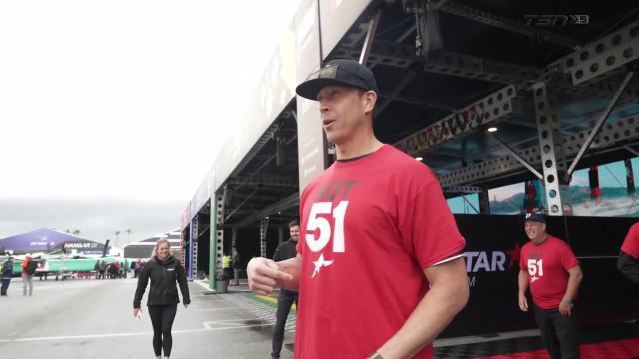 A man in a red "HOT 51" shirt gestures with his hands, talking to someone off-camera. A woman in a black jacket walks past, and the TSN5 logo is visible in the background.
