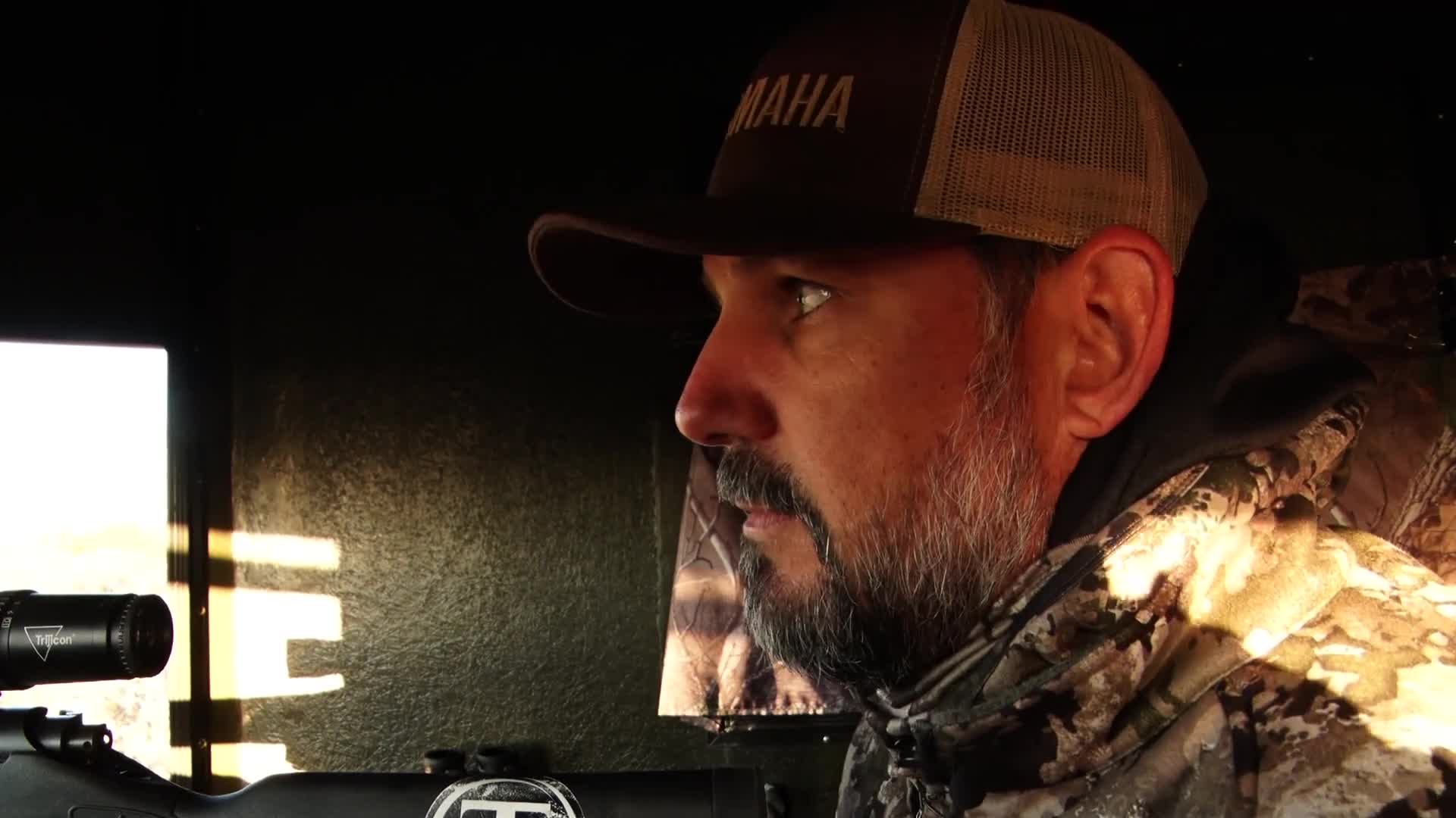 A man in a Yamaha hat and camouflage jacket peers intently from a blind. The rifle's scope is trained on something just outside the window.
