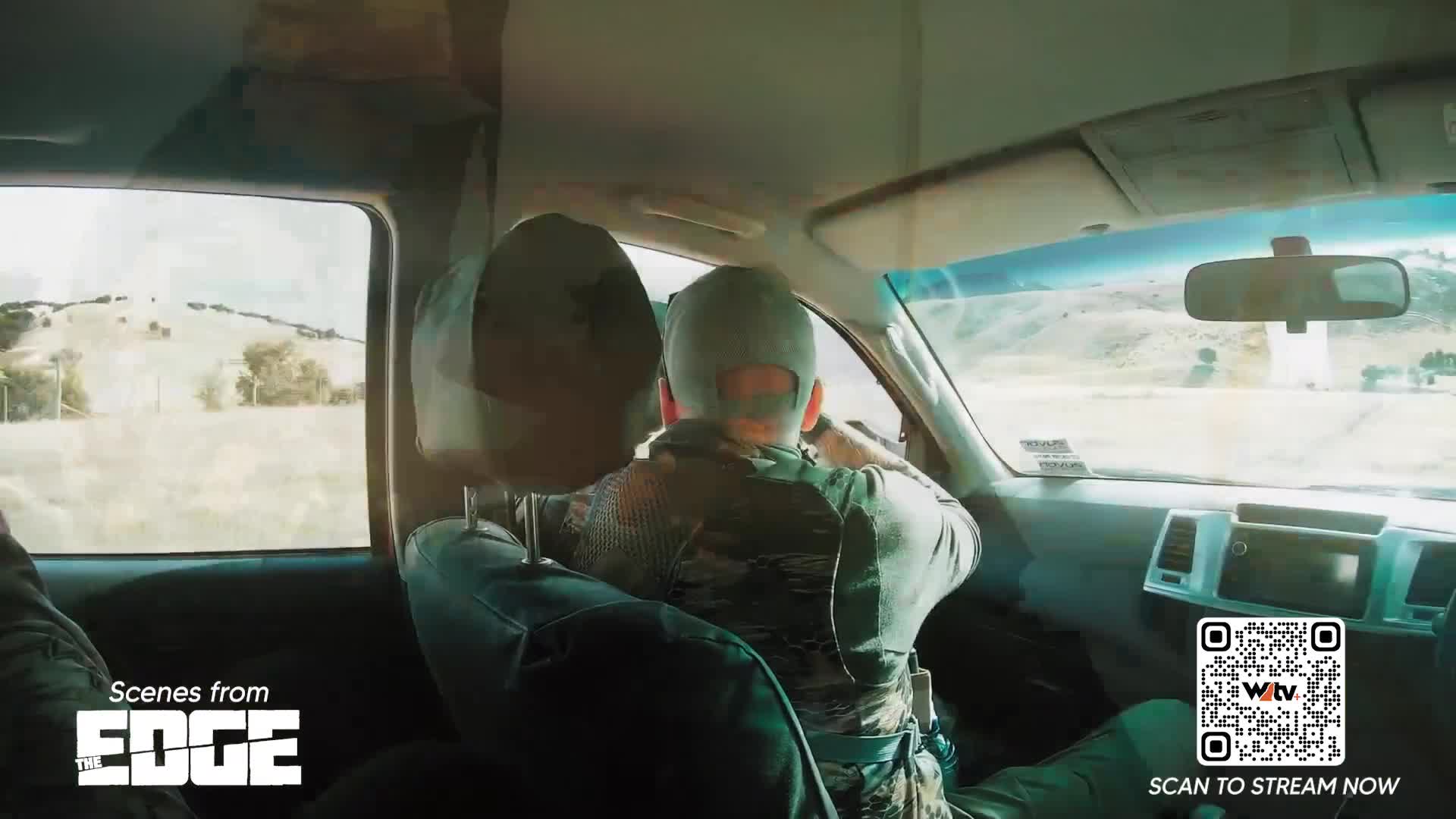The driver, wearing a cap and camouflage, looks out the window at the passing landscape. Through the vehicle's window, the hills of Canada's Wild TV are visible.
