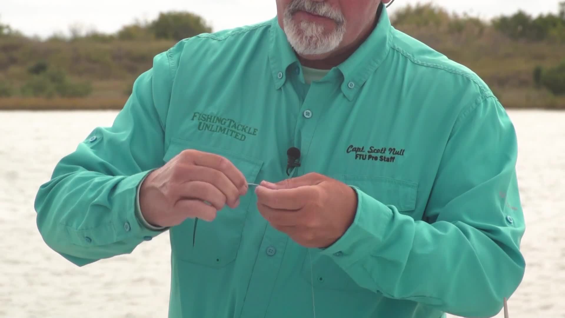 A man in a teal shirt carefully manipulates a thin fishing line between his fingers. He's tying a knot, the line looping and crossing as he works.
